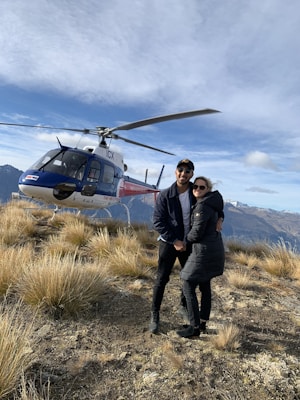 A couple is standing on a grassy, rocky hilltop with mountains in the background. They are dressed warmly, suggesting a cool climate. A helicopter is parked nearby, indicating adventure or travel.