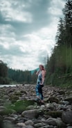 A woman stands confidently on a rocky riverbed surrounded by lush green forest and under a cloud-filled sky. She is wearing colorful athletic leggings and a matching top. The scene conveys a sense of tranquility and connection with nature.