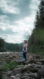 A woman stands confidently on a rocky riverbed surrounded by lush green forest and under a cloud-filled sky. She is wearing colorful athletic leggings and a matching top. The scene conveys a sense of tranquility and connection with nature.