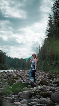 A woman stands confidently on a rocky riverbed surrounded by lush green forest and under a cloud-filled sky. She is wearing colorful athletic leggings and a matching top. The scene conveys a sense of tranquility and connection with nature.