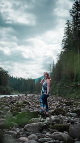 A woman stands confidently on a rocky riverbed surrounded by lush green forest and under a cloud-filled sky. She is wearing colorful athletic leggings and a matching top. The scene conveys a sense of tranquility and connection with nature.