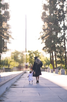 A woman wearing a graduation gown and cap walks hand in hand with a young child along a sunlit path. Tall trees line one side, with a reflective surface on the other side of the path.