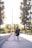 A woman wearing a graduation gown and cap walks hand in hand with a young child along a sunlit path. Tall trees line one side, with a reflective surface on the other side of the path.