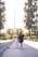 A woman wearing a graduation gown and cap walks hand in hand with a young child along a sunlit path. Tall trees line one side, with a reflective surface on the other side of the path.