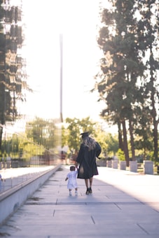 A woman wearing a graduation gown and cap walks hand in hand with a young child along a sunlit path. Tall trees line one side, with a reflective surface on the other side of the path.