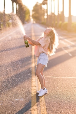 woman in white shirt and blue denim shorts holding green bottle standing on road during daytime