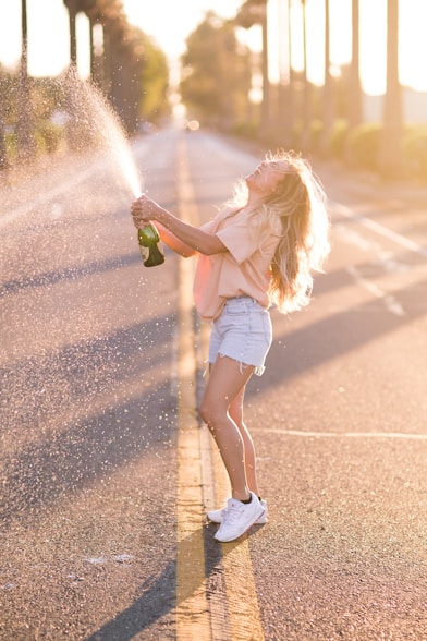 woman in white shirt and blue denim shorts holding green bottle standing on road during daytime
