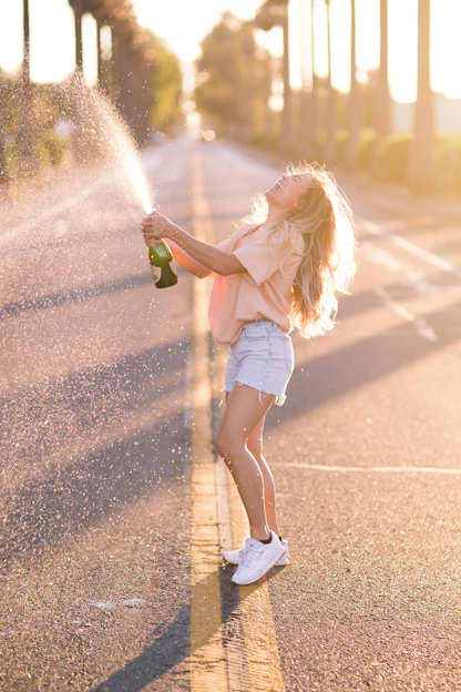 woman in white shirt and blue denim shorts holding green bottle standing on road during daytime