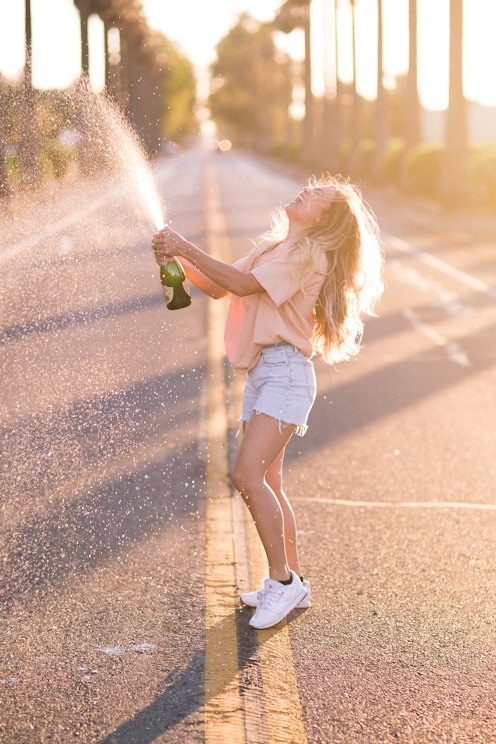 woman in white shirt and blue denim shorts holding green bottle standing on road during daytime