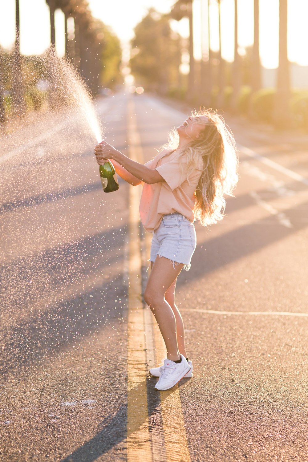 Mujer con camisa blanca y pantalones cortos de mezclilla azul sosteniendo una botella verde de pie en la carretera durante el día