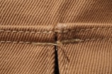 Detailed close-up view of brown fabric showcasing a seam with intricate stitching patterns. The texture is visible, with a clear focus on the woven fibers and stitch details.