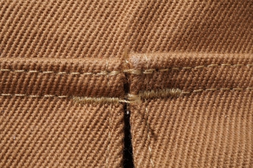 Detailed close-up view of brown fabric showcasing a seam with intricate stitching patterns. The texture is visible, with a clear focus on the woven fibers and stitch details.