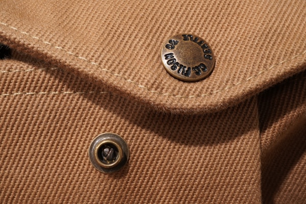 Close-up view of a textured brown fabric with visible stitching and two metallic buttons. One button has engraved text around its edge.