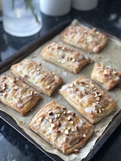 A baking tray filled with freshly baked pastries, likely puff pastries topped with chopped nuts and drizzled icing. They are arranged neatly on parchment paper and have a golden-brown crust.