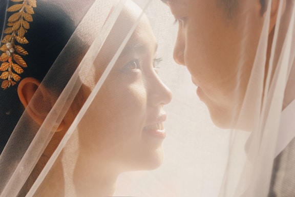 A close-up of a bride and groom gently facing each other under a soft veil, capturing a tender moment. The bride has golden leaf decorations in her hair.