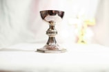 A silver chalice stands on a white surface, crafted with ornate detailing around its stem and base, reflecting light. In the background, a blurred golden cross can be seen.