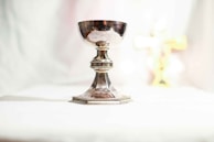 A silver chalice stands on a white surface, crafted with ornate detailing around its stem and base, reflecting light. In the background, a blurred golden cross can be seen.