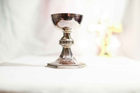 A silver chalice stands on a white surface, crafted with ornate detailing around its stem and base, reflecting light. In the background, a blurred golden cross can be seen.