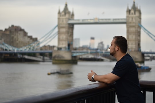 man in black shirt sitting on black wooden bench near bridge during daytime in Tower Bridge United Kingdom