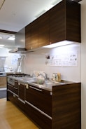 A modern kitchen setup featuring dark wood cabinetry with a glossy finish. The kitchen includes a stainless steel double sink and a gas stove with a steel hood. Under-cabinet lighting highlights the sleek and minimalistic design. Posters and informational sheets are displayed on the wall behind the sink. The flooring is made of light wood with a smooth finish.