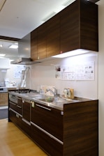 A modern kitchen setup featuring dark wood cabinetry with a glossy finish. The kitchen includes a stainless steel double sink and a gas stove with a steel hood. Under-cabinet lighting highlights the sleek and minimalistic design. Posters and informational sheets are displayed on the wall behind the sink. The flooring is made of light wood with a smooth finish.