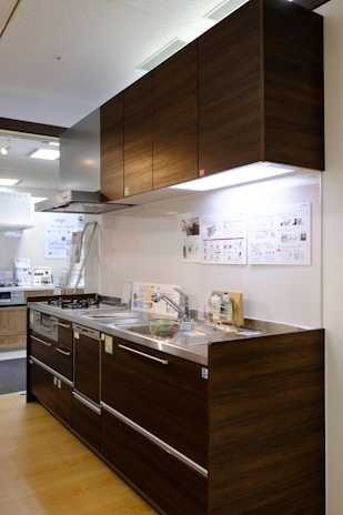 A modern kitchen setup featuring dark wood cabinetry with a glossy finish. The kitchen includes a stainless steel double sink and a gas stove with a steel hood. Under-cabinet lighting highlights the sleek and minimalistic design. Posters and informational sheets are displayed on the wall behind the sink. The flooring is made of light wood with a smooth finish.