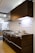 A modern kitchen setup featuring dark wood cabinetry with a glossy finish. The kitchen includes a stainless steel double sink and a gas stove with a steel hood. Under-cabinet lighting highlights the sleek and minimalistic design. Posters and informational sheets are displayed on the wall behind the sink. The flooring is made of light wood with a smooth finish.