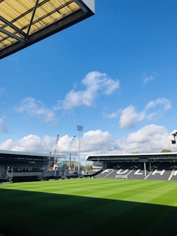 A football stadium with freshly cut green grass under a clear blue sky. There are two main stands visible, one with the word 'FULHAM' printed on the seats. Construction cranes are seen in the background, indicating ongoing development work.