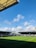 A football stadium with freshly cut green grass under a clear blue sky. There are two main stands visible, one with the word 'FULHAM' printed on the seats. Construction cranes are seen in the background, indicating ongoing development work.