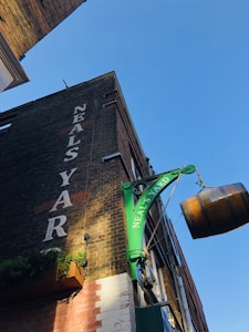 An upward view of a brick building corner with a large, green hanging sign that reads 'Neal’s Yard'. A wooden barrel sign is also hanging from the structure. The building facade has 'NEAL'S YARD' painted vertically in large white letters. Part of the building base is painted white with some vegetation.