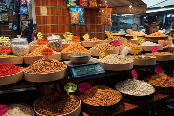 Various types of nuts and seeds are displayed in round wooden trays at a market stall. The trays are filled with pistachios, almonds, sunflower seeds, and other snacks. Handwritten signs in a foreign language indicate the names or prices. Clear glass jars filled with colorful candies are visible, and a digital scale is on the counter. The market setting is busy, with shoppers in the background.