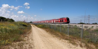 A long red passenger train moves along railway tracks elevated on a gravel embankment. The train is surrounded by a rural landscape with a dirt path in the foreground and green fields with wildflowers. A wire fence runs parallel to the train tracks, and several power lines and poles are visible in the background under a blue sky with scattered clouds.