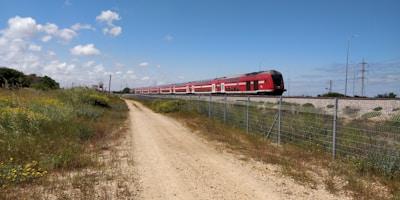A long red passenger train moves along railway tracks elevated on a gravel embankment. The train is surrounded by a rural landscape with a dirt path in the foreground and green fields with wildflowers. A wire fence runs parallel to the train tracks, and several power lines and poles are visible in the background under a blue sky with scattered clouds.