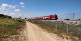 A long red passenger train moves along railway tracks elevated on a gravel embankment. The train is surrounded by a rural landscape with a dirt path in the foreground and green fields with wildflowers. A wire fence runs parallel to the train tracks, and several power lines and poles are visible in the background under a blue sky with scattered clouds.