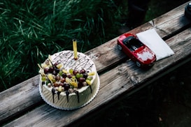 A round cake decorated with a variety of fruits such as grapes and pineapple slices sits on a rustic wooden bench. The cake is topped with a decorative sparkler candle. Next to the cake, there is a small red toy car and a folded white napkin.