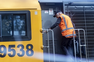 man in orange jacket and black pants standing in front of yellow train