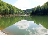 A serene lake surrounded by lush green forested hills, with a clear reflection of the sky and clouds on the water's surface. The scene is tranquil and picturesque, capturing the harmony of nature.