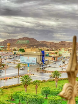 A scenic cityscape featuring a prominent road intersection surrounded by vibrant green trees and well-maintained landscaping. The skyline includes an array of buildings, a significant billboard with text, and a large flag design laid out on a hillside. In the background, there are mountains under a cloudy sky, creating a dramatic and picturesque atmosphere.