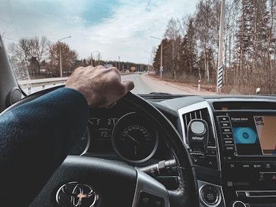 A person is driving a car on an open road, with a view of the dashboard and steering wheel. The steering wheel has a brand logo, and the driver's hand is visible gripping it. A road flanked by trees extends into the distance under a partly cloudy sky.