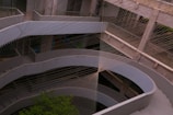 A multi-story concrete structure with curved architectural elements is intersected by a complex network of intersecting strings or cables, creating an artistic geometric pattern. The setting appears modern and industrial, with the presence of some greenery visible in the lower section.
