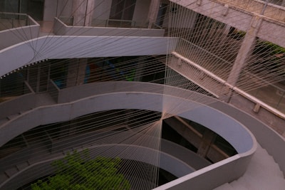 A multi-story concrete structure with curved architectural elements is intersected by a complex network of intersecting strings or cables, creating an artistic geometric pattern. The setting appears modern and industrial, with the presence of some greenery visible in the lower section.