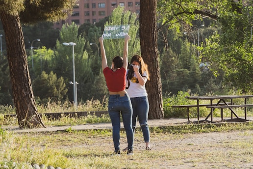 Two people are standing in a park. One is holding up a transparent sign with text that appears reversed. The other is standing beside them with a hand near their face. There are trees in the background, and a wooden picnic table is visible.