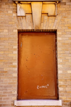 A closed window or doorway with a rust-colored metal covering set into a light brown brick wall. Above the window is a decorative stone lintel. The metal surface is weathered and shows signs of peeling paint. Chalk writing of the equation e=mc² is visible at the bottom of the panel.