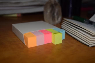 A stack of multicolored sticky notes is arranged neatly on a wooden surface. The colors include orange, pink, yellow, lime green, and light blue. Adjacent to the sticky notes is a notepad with a few pages open and a pen resting on top. Background elements include a blurry stack of papers and a stone-like object.