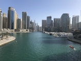A busy waterfront cityscape featuring modern high-rise buildings flanking both sides of a canal with turquoise water. Numerous yachts are docked along the marina, and a small boat with several people is visible on the right side of the canal.