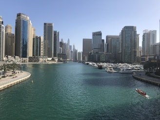 A busy waterfront cityscape featuring modern high-rise buildings flanking both sides of a canal with turquoise water. Numerous yachts are docked along the marina, and a small boat with several people is visible on the right side of the canal.