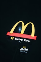 A brightly lit McDonald's drive-thru sign is prominently displayed against a dark background. The iconic yellow 'M' arches are situated atop a red and black base with the words 'Drive Thru' in white, accompanied by the McCafé logo below.