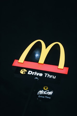 A brightly lit McDonald's drive-thru sign is prominently displayed against a dark background. The iconic yellow 'M' arches are situated atop a red and black base with the words 'Drive Thru' in white, accompanied by the McCafé logo below.