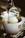 Coffee is being poured over a scoop of vanilla ice cream in a white cup with a red 'illy' logo. The setup rests on a metal surface, possibly a counter or drip tray.