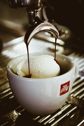 Coffee is being poured over a scoop of vanilla ice cream in a white cup with a red 'illy' logo. The setup rests on a metal surface, possibly a counter or drip tray.